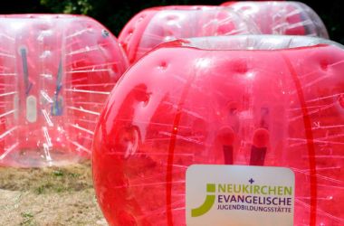 Rötliche Bumper-Balls mit dem JuBi Neukirchen Logo für das Spiel Bubble Soccer auf von Sonne beschienener Wiese.