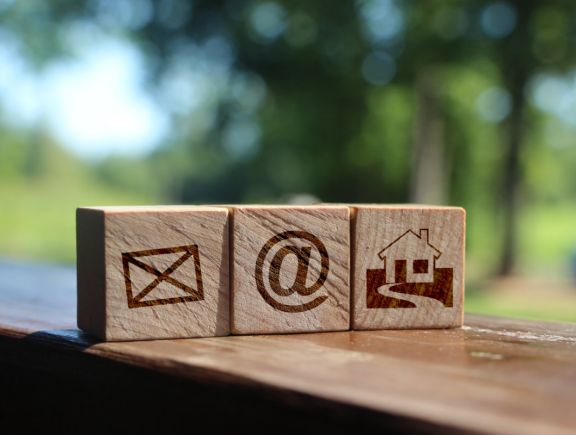 Drei Holzwürfel mit Symbolen für Brief, @-Zeichen und Anfahrt auf einem Holzbrett mit unscharfen grünen Bäumen im Hintergrund