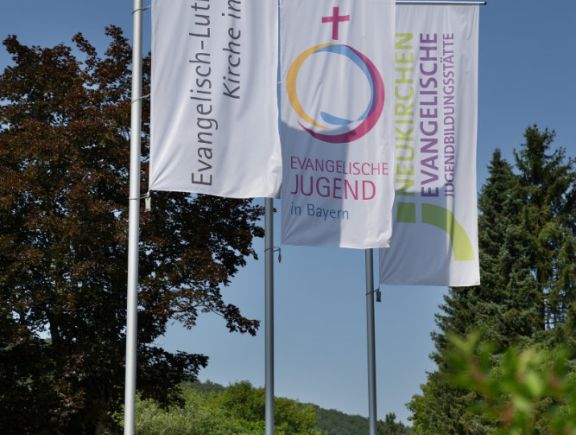 Große Fahnen an Mästen mit den Logos von der Evangelisch-Lutherischen Kirche in Bayer, der Evangelischen Jugend in Bayern und der Evangelischen Jugendbildungsstätte Neukirchen
