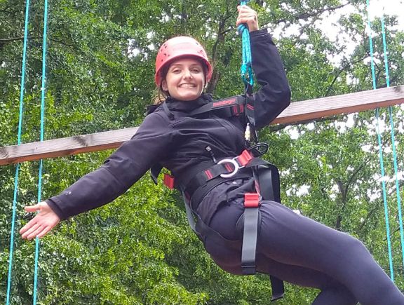 Frau in Sicherungsseil im Klettergarten hängend, posiert und grinst dabei freundlich